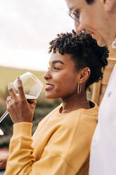 Side view of man embracing black woman standing with glass of wine on balcony while enjoying weekend and looking away - ADSF25441