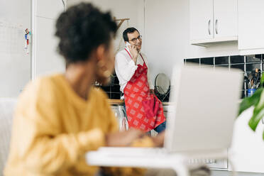 Mann in Schürze, der in der Küche mit seinem Smartphone spricht, und schwarze Frau, die zu Hause auf ihrem Laptop surft - ADSF25443