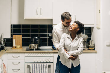 Entzückter Mann umarmt lächelnde schwarze Frau von hinten, während sie in der Küche stehen und sich gegenseitig anschauen - ADSF25457