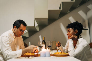 Seitenansicht der glücklichen multirassischen Paar sitzt am Tisch mit Kerzen und Wein beim Essen köstliche Gerichte während romantisches Abendessen zu Hause - ADSF25459