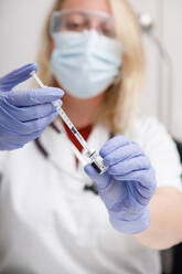 Blurred unrecognizable medic in protective face shield mask and latex gloves with vial of coronavirus vaccine and syringe showing to camera while standing in hospital room - ADSF25470