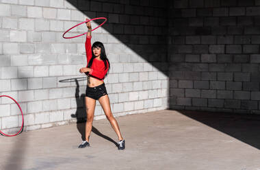 Junge tätowierte Frau in Activewear wirbelt Hula-Hoop-Reifen, während sie gegen Backsteinwände mit Schatten tanzt und im Sonnenlicht nach vorne schaut - ADSF25482