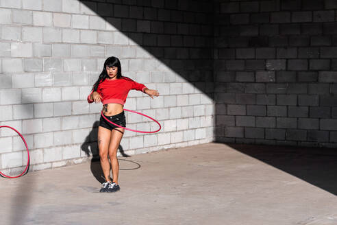 Junge tätowierte Frau in Activewear wirbelt Hula-Hoop-Reifen beim Tanzen gegen Backsteinwände mit Schatten - ADSF25483