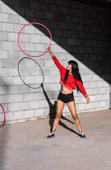Young tattooed woman in activewear twirling hula hoop while dancing against brick walls with shadows and looking forward in sunlight - ADSF25485
