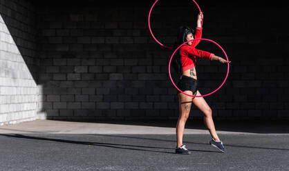 Junge tätowierte Frau in Activewear wirbelt Hula-Hoop-Reifen, während sie gegen Backsteinwände mit Schatten tanzt und im Sonnenlicht nach vorne schaut - ADSF25486