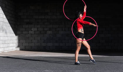 Young tattooed woman in activewear twirling hula hoop while dancing against brick walls with shadows and looking forward in sunlight - ADSF25486