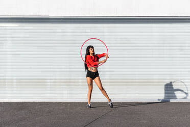 Junge tätowierte Frau in Activewear wirbelt Hula-Hoop-Reifen beim Tanzen gegen Backsteinwände mit Schatten - ADSF25487