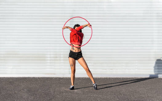 Junge tätowierte Frau in Activewear wirbelt Hula-Hoop-Reifen, während sie gegen Backsteinwände mit Schatten tanzt und im Sonnenlicht nach vorne schaut - ADSF25488