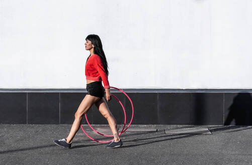 Seitenansicht einer fitten jungen Frau in Sportkleidung mit Reifen, die auf einem Gehweg spazieren geht und im Sonnenlicht nach vorne schaut - ADSF25491