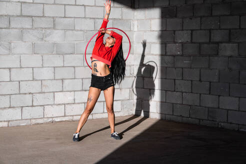 Junge tätowierte Frau in Activewear wirbelt Hula-Hoop-Reifen, während sie gegen Backsteinwände mit Schatten tanzt und im Sonnenlicht nach vorne schaut - ADSF25494