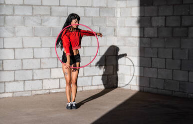 Young tattooed woman in activewear twirling hula hoop while dancing against brick walls with shadows - ADSF25495