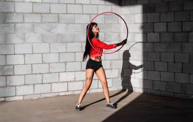 Junge tätowierte Frau in Activewear wirbelt Hula-Hoop-Reifen, während sie gegen Backsteinwände mit Schatten tanzt und im Sonnenlicht nach vorne schaut - ADSF25496