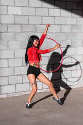 Junge tätowierte Frau in Activewear wirbelt Hula-Hoop-Reifen, während sie gegen Backsteinwände mit Schatten tanzt und im Sonnenlicht nach vorne schaut - ADSF25497