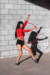Young tattooed woman in activewear twirling hula hoop while dancing against brick walls with shadows and looking forward in sunlight - ADSF25497