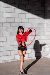 Young tattooed woman in activewear twirling hula hoop while dancing against brick walls with shadows - ADSF25499