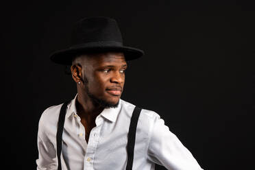 Young masculine ethnic male model in hat and trousers standing while looking away on black background with smoke - ADSF25513