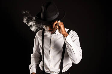 Männlicher Afroamerikaner in weißem Hemd und Hut, der beim Rauchen einer E-Zigarette Dampf ausstößt - ADSF25521