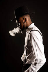 Side view of masculine African American male in white shirt and hat exhaling vapor while smoking e cigarette - ADSF25522