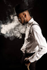 Side view of masculine African American male in white shirt and hat exhaling vapor while smoking e cigarette - ADSF25523
