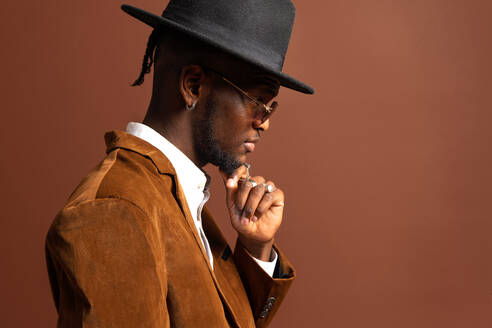 Seitenansicht eines jungen Afroamerikaners in modischer Kleidung und Hut, der auf braunem Hintergrund wegschaut - ADSF25529