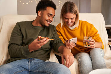 Zufriedenes gemischtrassiges Paar sitzt im Wohnzimmer, isst Pizza und sieht sich am Wochenende ein lustiges Video auf dem Smartphone an - ADSF25536