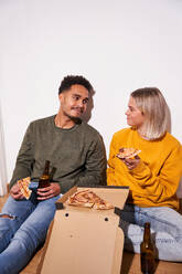 Cheerful multiethnic couple sitting on floor at home while eating delicious pizza and drinking beer together - ADSF25540