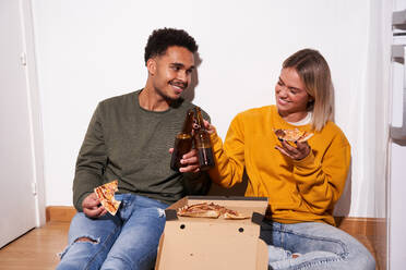 Fröhliches multiethnisches Paar, das zu Hause auf dem Boden sitzt, während es leckere Pizza isst und gemeinsam Bier trinkt - ADSF25541