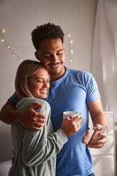 Side view of smiling multiracial couple in pajamas hugging in morning while taking selfie in morning at home - ADSF25544