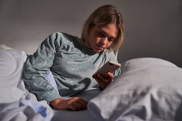 Gelassene Frau liegt im Bett unter einer Decke und surft im Internet auf ihrem Mobiltelefon nach dem Aufwachen am Morgen - ADSF25546