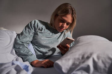 Serene female lying in bed under blanket and surfing Internet on mobile phone after awakening in morning - ADSF25546