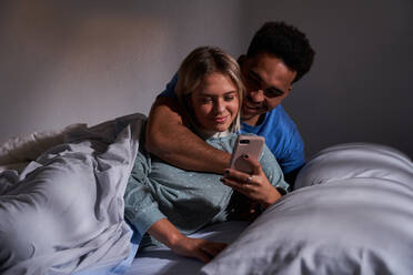 Erfreuliches gemischtrassiges Paar, das auf dem Bett liegt, während es auf dem Smartphone surft und gemeinsam Spaß am Morgen hat - ADSF25547