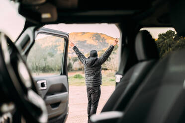 Rückansicht durch das Innere eines Autos eines Mannes, der bei Sonnenaufgang mit erhobenen Armen als Zeichen der Freiheit die Berglandschaft betrachtet - ADSF25558