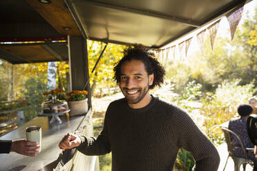 Porträt eines glücklichen männlichen Kunden, der an einem Imbisswagen im Park Kaffee bestellt - CAIF31330