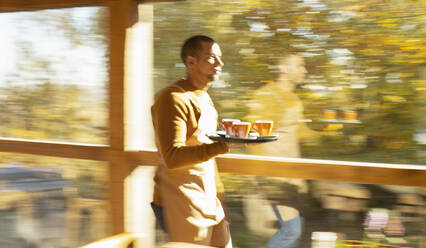 Männlicher Barista trägt Tablett mit Kaffee am Fenster eines sonnigen Herbstcafés entlang - CAIF31349