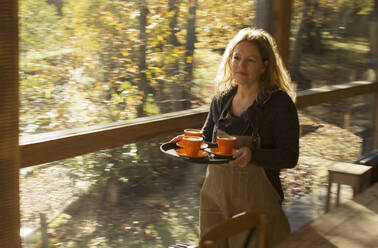 Weiblicher Barista trägt Kaffee auf einem Tablett am Fenster eines sonnigen Herbstcafés entlang - CAIF31355