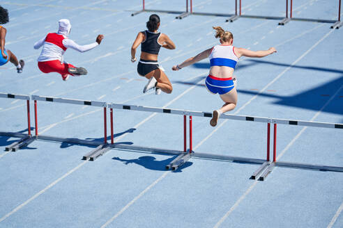 Female track and field athletes jumping hurdles on sunny track - CAIF31395
