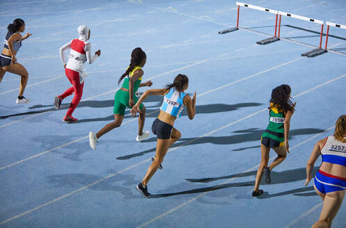 Female track and field athletes running hurdle race on blue track - CAIF31403