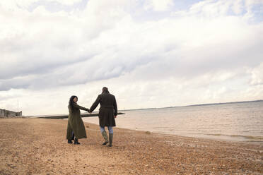 Couple in winter coats holding hands walking on ocean beach - CAIF31585