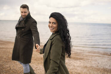 Portrait happy couple in winter coats holding hands on ocean beach - CAIF31597