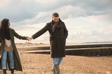 Paar in Wintermänteln, das sich an den Händen hält und am sonnigen Strand spazieren geht - CAIF31605