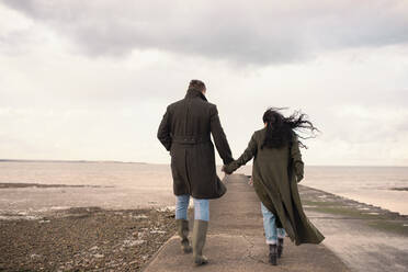 Couple in winter coats holding hands on ocean beach jetty - CAIF31612