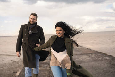 Happy couple in winter coats holding hands on ocean jetty - CAIF31618