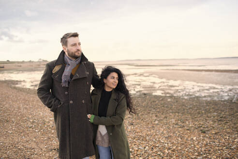 Affectionate couple in winter coats walking on ocean beach - CAIF31621