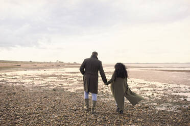 Paar in Wintermänteln, das sich an den Händen hält und am Strand spazieren geht - CAIF31632