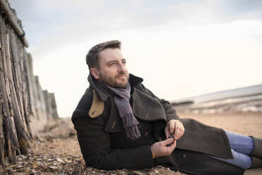 Serene man in winter coat relaxing on beach - CAIF31634