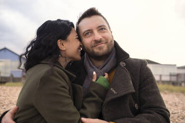 Happy affectionate couple in winter coats hugging on beach - CAIF31645