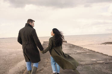 Couple in winter coats holding hands on ocean beach jetty - CAIF31657