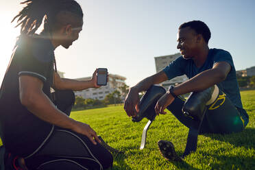 Trainer mit Smartphone im Gespräch mit amputiertem Athleten im sonnigen Gras - CAIF31675