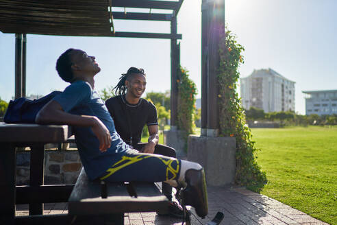 Männlicher amputierter Sportler und Trainer auf einer sonnigen Parkbank - CAIF31679