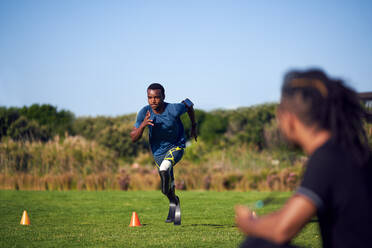 Coach watching young male amputee athlete running drills in park - CAIF31699